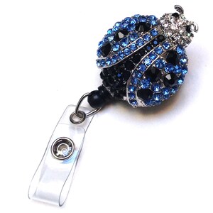 Peut inclure: Un porte-badge en plastique transparent avec un fermoir pivotant argent&eacute; et un charme en forme de coccinelle orn&eacute; de bijoux bleus et noirs.