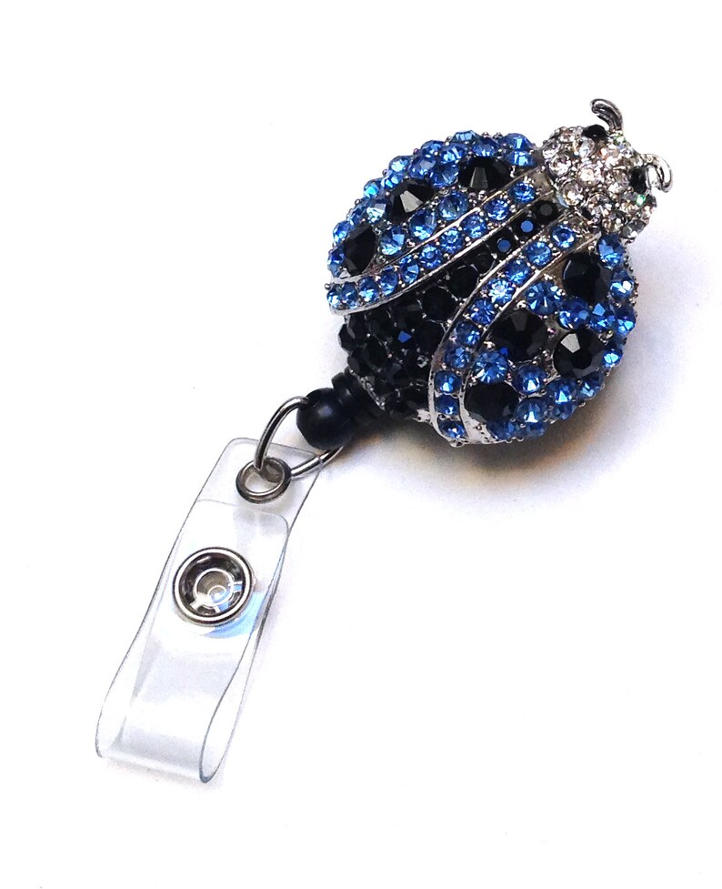 Peut inclure: Un porte-badge en plastique transparent avec un fermoir pivotant argent&eacute; et un charme en forme de coccinelle orn&eacute; de bijoux bleus et noirs.