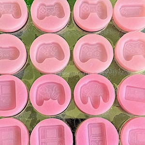 May include: A tray of pink, circular molds featuring video game controller and handheld console designs. The molds are arranged in a grid pattern, perfect for crafting themed items. The tray is clear and the background is a dark blue.