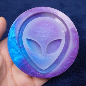 May include: A round, multi-colored alien-themed resin coaster. The coaster features a central alien head design in a light purple color, surrounded by a ring of purple and blue hues. The coaster is held in a hand.