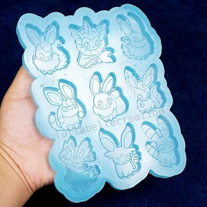 May include: A light blue silicone mold featuring nine cartoon bunny designs. The mold is held in a hand against a dark blue background. The designs are detailed with various poses and expressions.