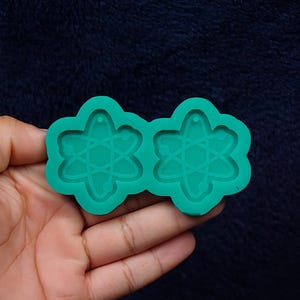 May include: A teal-colored silicone mold featuring two atom-shaped cavities. The mold is held in a hand against a dark blue background. Each cavity has a flower-like outline with an atom design inside.