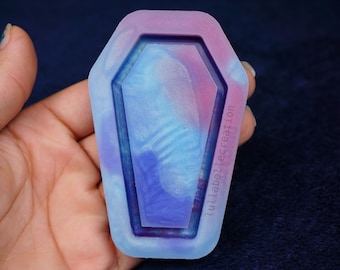 May include: A coffin-shaped resin piece with a marbled effect. The colors transition from light blue to purple and pink. The interior has a glossy, swirling appearance. The text "lullabellecreation" is visible on the side.
