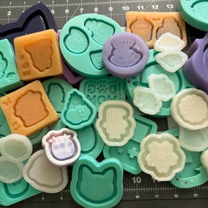 May include: A collection of colorful silicone molds in various shapes, including flowers, animals, and geometric designs. The molds are in shades of teal, orange, purple, and white. Some molds have detailed designs, and others are simple shapes.