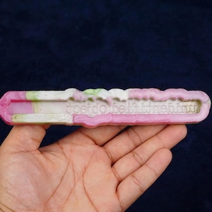 May include: A pink, white, and green rectangular object with the text "best fit itebinu" is held in a hand. The object has a wavy top edge and appears to be made of a solid material. The background is a dark blue.