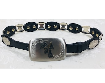 May include: A black leather belt with a silver buckle featuring a stylized cowboy riding a horse. The belt has silver oval-shaped links with studs.