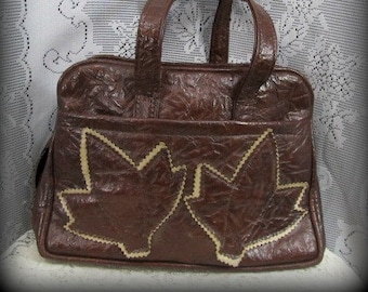 May include: A brown leather handbag with a top handle and a flap closure. The flap has two large leaf-shaped designs with a stitched edge.