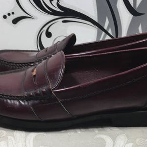 Puede incluir: Un par de mocasines de cuero burdeos con un dise&ntilde;o cl&aacute;sico de tira penny. Los zapatos tienen una punta ligeramente redondeada y una suela gruesa de goma negra.