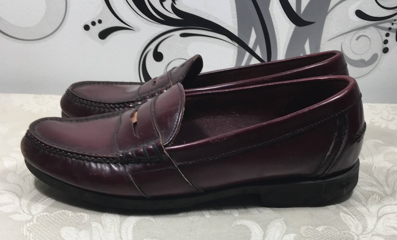 Puede incluir: Un par de mocasines de cuero burdeos con un dise&ntilde;o cl&aacute;sico de tira penny. Los zapatos tienen una punta ligeramente redondeada y una suela gruesa de goma negra.