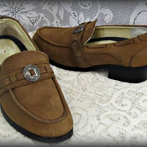 May include: A pair of brown leather loafers with a silver buckle detail. The loafers have a low heel and a rounded toe.