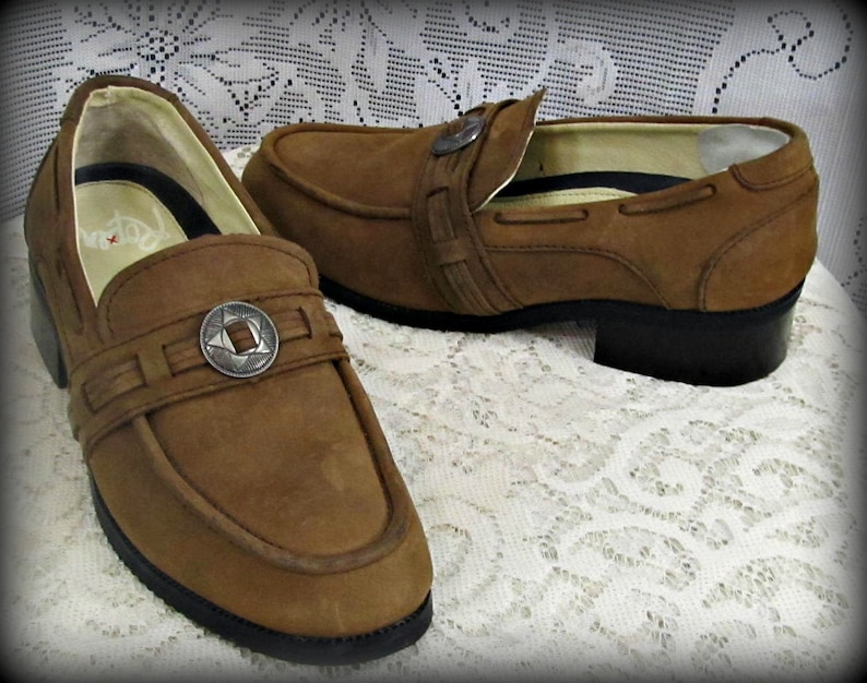 May include: A pair of brown leather loafers with a silver buckle detail. The loafers have a low heel and a rounded toe.
