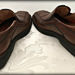 May include: A pair of brown leather slip-on shoes with black soles. The shoes have a stitched design and are worn.