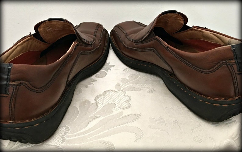 May include: A pair of brown leather slip-on shoes with black soles. The shoes have a stitched design and are worn.