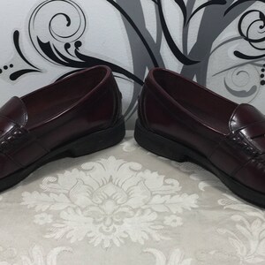 Puede incluir: Un par de mocasines de cuero burdeos con un acento dorado en la parte superior. Los mocasines tienen un estilo cl&aacute;sico de mocas&iacute;n penny.