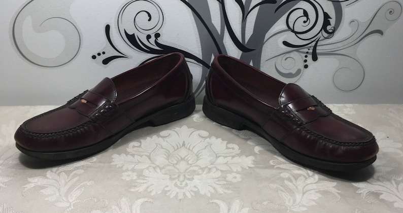 Puede incluir: Un par de mocasines de cuero burdeos con un acento dorado en la parte superior. Los mocasines tienen un estilo cl&aacute;sico de mocas&iacute;n penny.