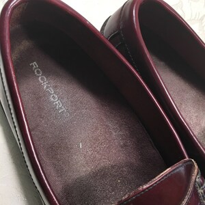 Puede incluir: Un par de mocasines de cuero burdeos con el nombre de la marca "Rockport" impreso en la plantilla.