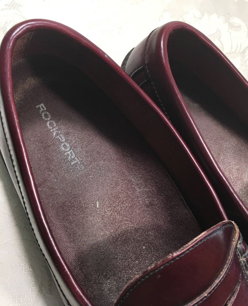 Puede incluir: Un par de mocasines de cuero burdeos con el nombre de la marca "Rockport" impreso en la plantilla.