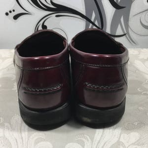 Puede incluir: Un par de mocasines de cuero burdeos con un acabado pulido. Los zapatos tienen una suela de goma negra y una costura cosida alrededor de la parte superior.