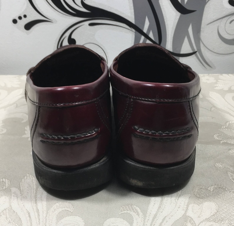 Puede incluir: Un par de mocasines de cuero burdeos con un acabado pulido. Los zapatos tienen una suela de goma negra y una costura cosida alrededor de la parte superior.