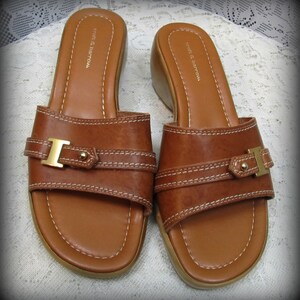 May include: A pair of brown leather slide sandals with a gold buckle and white stitching. The sandals have a low wedge heel and are labeled "croft & barrow".