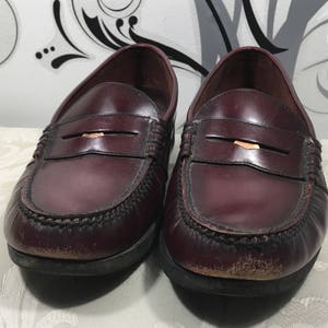 Puede incluir: Un par de mocasines de cuero burdeos con un detalle de tira de penique. Los zapatos tienen una suela marr&oacute;n oscuro y un aspecto ligeramente desgastado.