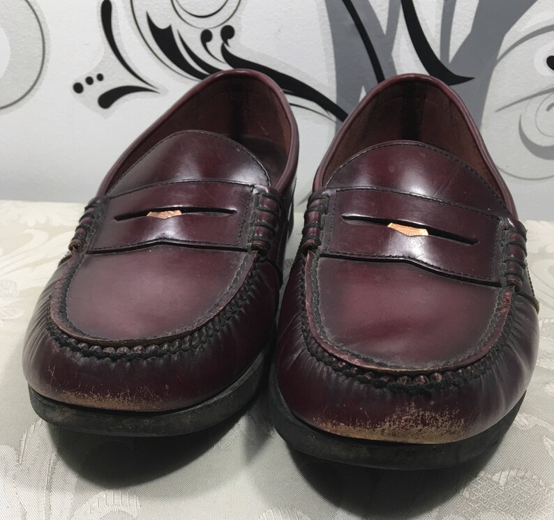 Puede incluir: Un par de mocasines de cuero burdeos con un detalle de tira de penique. Los zapatos tienen una suela marr&oacute;n oscuro y un aspecto ligeramente desgastado.