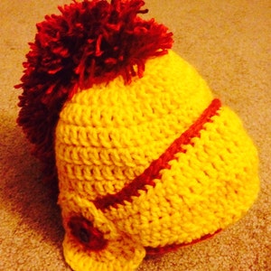 May include: A yellow crocheted hat with a red pom-pom on top and a red stripe around the brim.