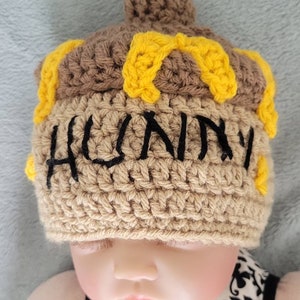 May include: A brown crocheted hat with a yellow crocheted design and the word "HUNNY" embroidered in black. The hat is worn by a baby.