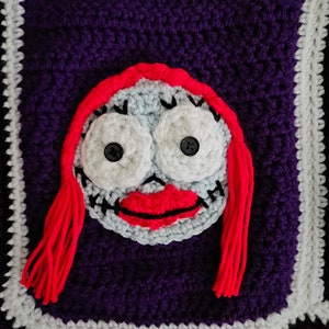 May include: Crocheted square with a purple background and a white border. The center of the square features a red-haired character with a white face, black eyes, and a red mouth.
