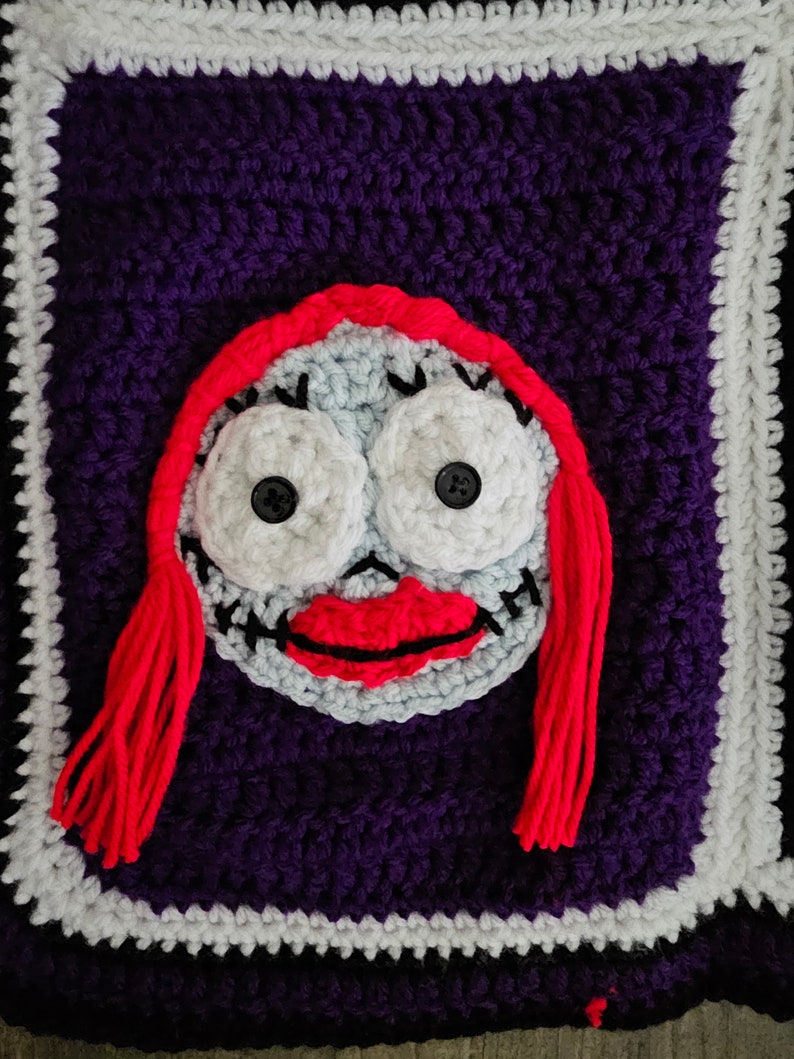 May include: Crocheted square with a purple background and a white border. The center of the square features a red-haired character with a white face, black eyes, and a red mouth.