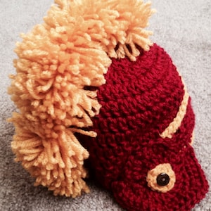 May include: A crocheted hat with a red base and a yellow pom-pom on top. The hat is shaped like a helmet with a black button on the front.