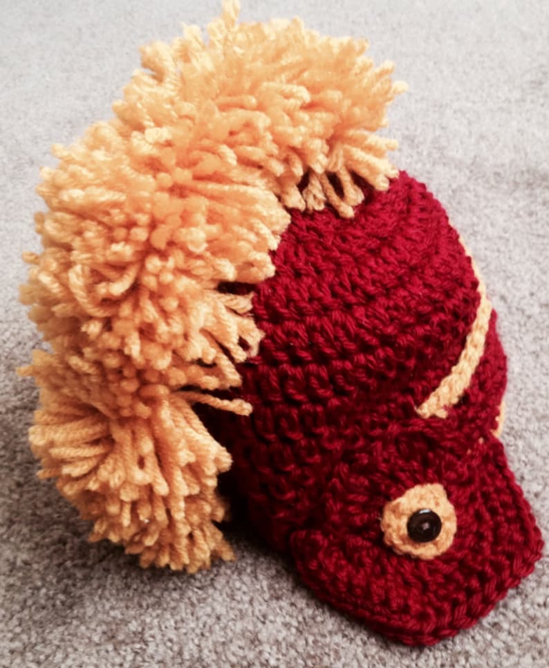 May include: A crocheted hat with a red base and a yellow pom-pom on top. The hat is shaped like a helmet with a black button on the front.