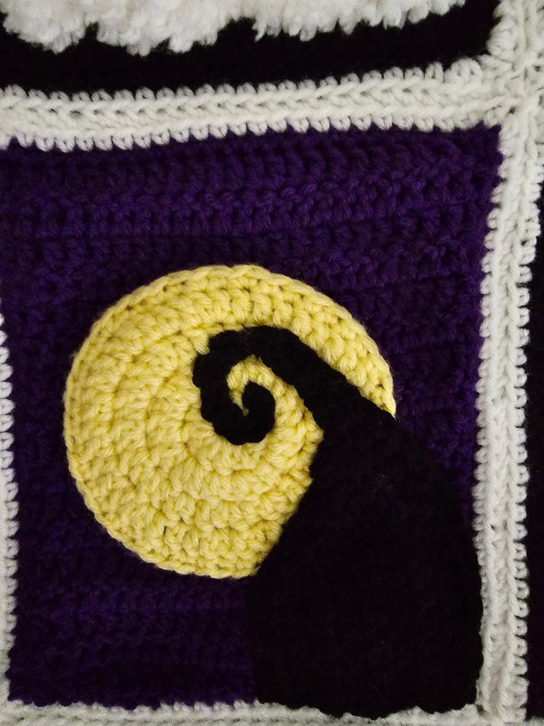 May include: A crocheted square with a purple background, a yellow crescent moon, and a black silhouette of a spiral. The square is bordered with white crochet.