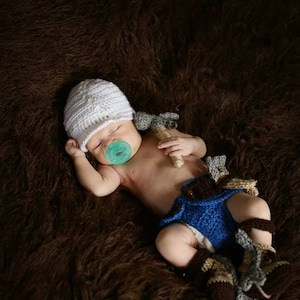 May include: A newborn baby wearing a white crocheted hat, a blue crocheted diaper, and a pacifier. The baby is lying on a brown fuzzy blanket and holding a crocheted toy.