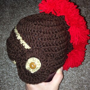 May include: A brown crocheted hat with a large red pom-pom on top. The hat has a brown button on the front.