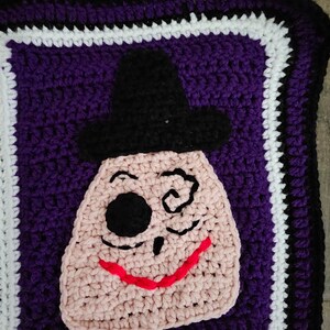 May include: Crocheted square with a purple background and a white and black border. The center of the square features a pink crocheted face with a black eye patch, a black hat, and a red smile.