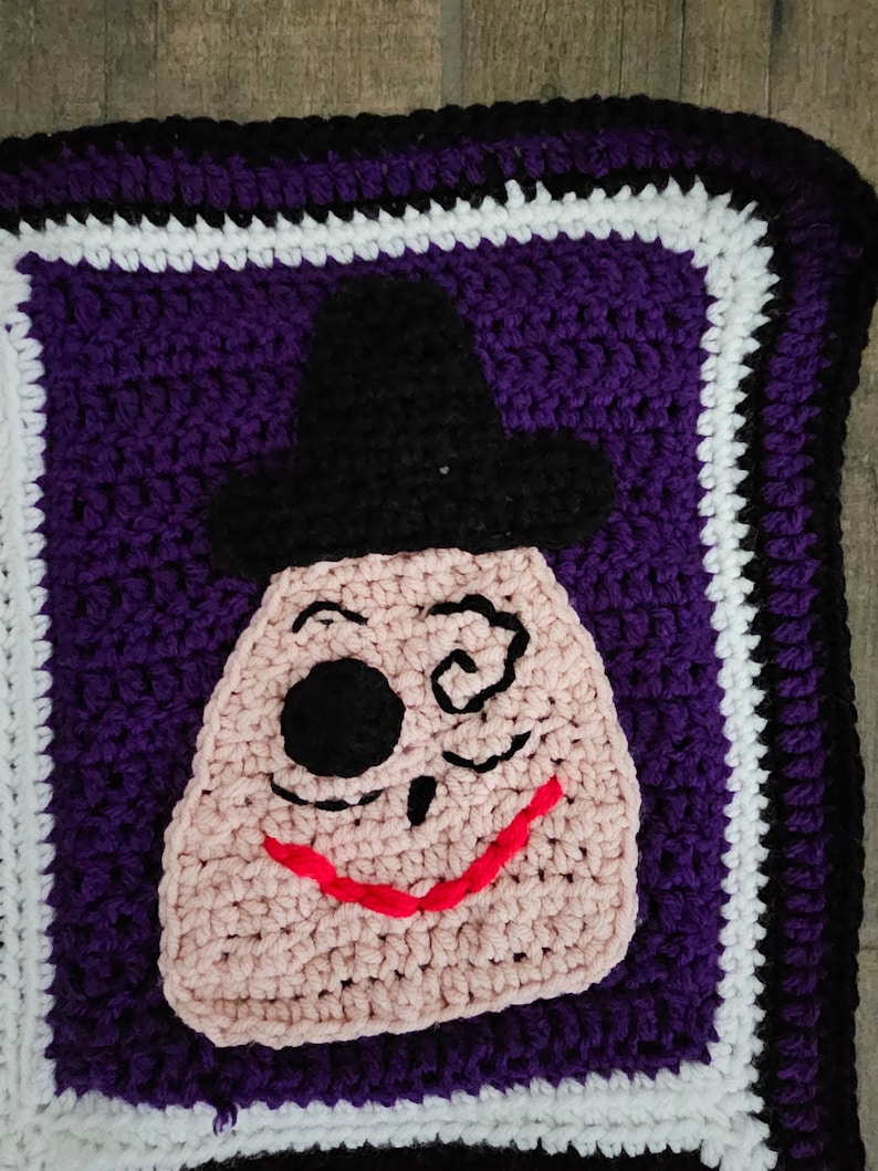 May include: Crocheted square with a purple background and a white and black border. The center of the square features a pink crocheted face with a black eye patch, a black hat, and a red smile.