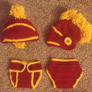 May include: A crocheted baby outfit in maroon and yellow. The outfit includes a hat, a diaper cover, and a helmet. The hat has a yellow pom-pom on top. The helmet has a yellow pom-pom on top and a yellow stripe around the bottom.