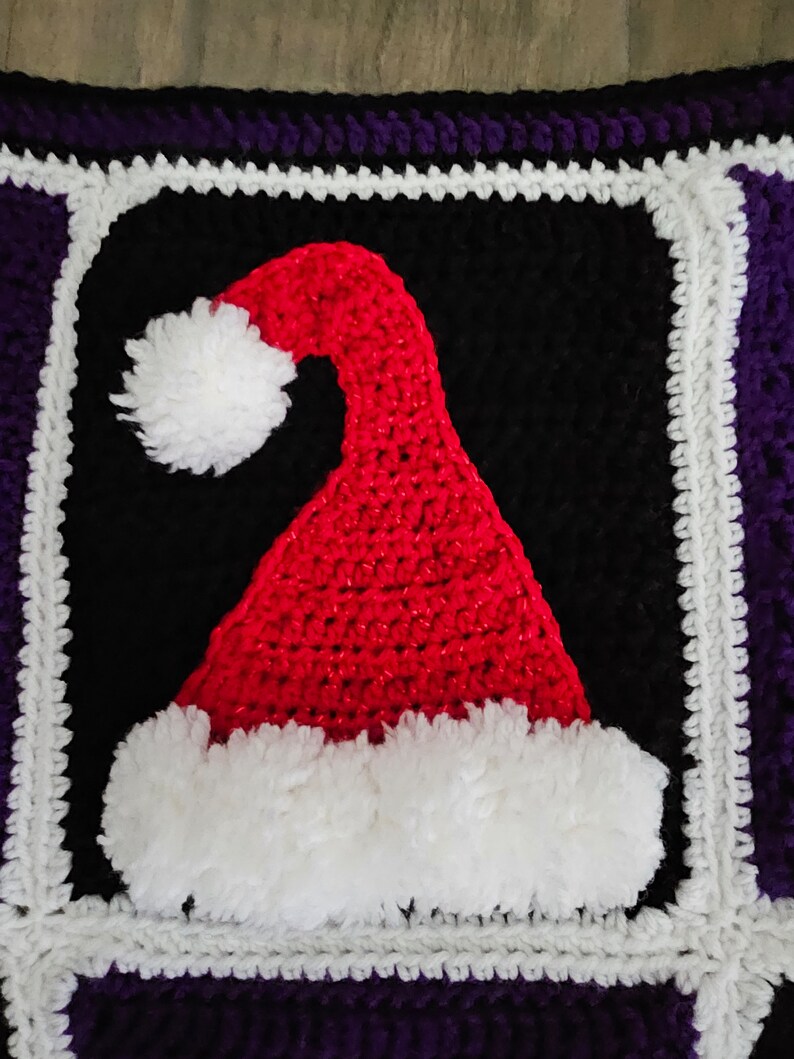 May include: A crocheted square with a red and white Santa hat design. The hat is surrounded by a black square with a white border and a purple border.