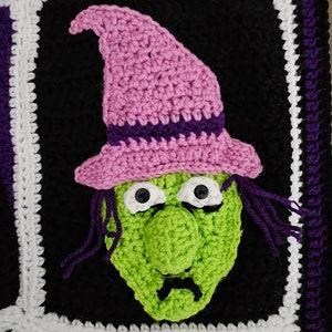 May include: Crocheted witch face with a pink hat, green face, and black eyes. The witch face is framed by a black, white, and purple border.
