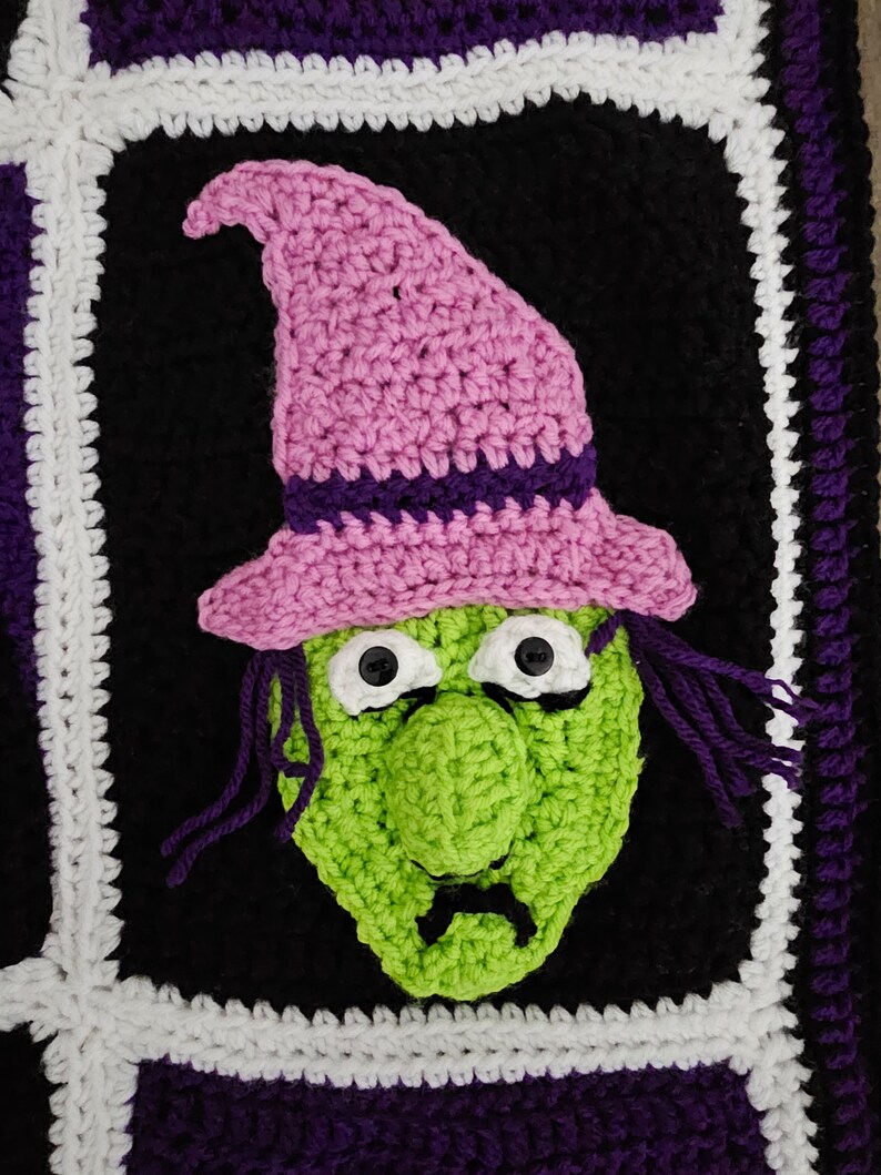 May include: Crocheted witch face with a pink hat, green face, and black eyes. The witch face is framed by a black, white, and purple border.