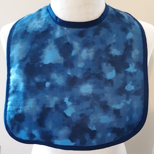 May include: A blue and white tie-dye bib with a dark blue trim. The bib is made of a soft, comfortable fabric and is perfect for protecting clothes from spills and messes.