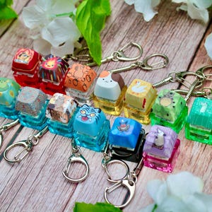 May include: A collection of colorful keycap keychains with various designs, including animals and patterns. Each keycap is mounted on a clear, rectangular base and attached to a silver keychain. The keycaps are red, orange, blue, green, and purple.