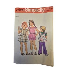 Può includere: Una busta per cartamodello Simplicity 7459 con illustrazioni di tre bambini che indossano abiti diversi. I modelli includono un vestito a quadri, un top rosa con pantaloncini e un top a stampa floreale con pantaloni viola. Il testo riporta 7459, Simplicity, Size 3.