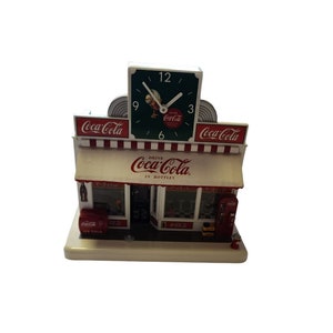 Puede incluir: Un reloj con temática de Coca-Cola con forma de edificio de tienda vintage. La esfera del reloj es verde con números y manecillas blancas. La tienda tiene un toldo rojo y blanco con las palabras "Drink Coca-Cola in Bottles".
