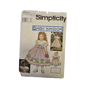 Może przedstawiać: Koperta z wykrojem krawieckim Simplicity Daisy Kingdom 9092 dla rozmiarów AA 3, 4, 5, 6. Biała papierowa koperta zawiera ilustracje sukienki z różową plisowaną spódnicą, niebieską kokardą i kwiatowym obszyciem oraz wzór na sukienkę dla lalki.
