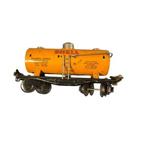 May include: Vintage Lionel Lines G55 Shell tank car. The toy train car is painted orange with black wheels and undercarriage. The words "SHELL" and "LIONEL LINES" are visible on the tank car. The car has a ladder and other details.