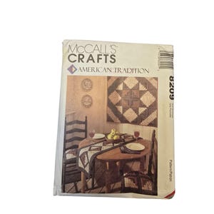 Puede incluir: Un sobre de patrón de McCall's Crafts con una escena rústica con una mesa redonda de madera para dos personas. La escena incluye un edredón, un camino de mesa y cojines de silla con un diseño geométrico. El texto "American Tradition" es visible.