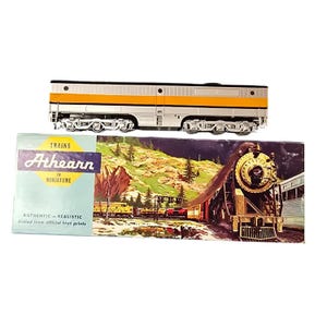 May include: Athearn model train set featuring a silver and yellow passenger car and a box with a train illustration. The box reads "Trains in Miniature" and "Authentic - Realistic". The passenger car has black wheels and is on a white background.