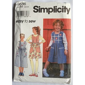Może przedstawiać: Wzór szycia Simplicity numer 9636, rozmiar BB 5, 6, 7, 8. Wzór zawiera instrukcje do uszycia trzech różnych sukienek dla dziewczynek. Sukienki są w stylu casual i mają kieszenie. Pierwsza sukienka to niebiesko-biała w paski sukienka z czerwonymi sercami. Druga sukienka to sukienka w kwiatowy wzór z czerwoną i białą kratą w postaci fartucha. Trzecia sukienka to niebieska dżinsowa sukienka z białymi haftowanymi truskawkami.